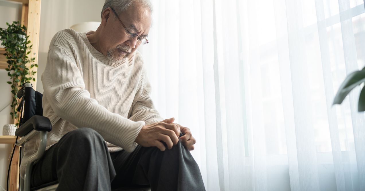 Elderly man in a wheelchair holding his knee with a pained expression indoors