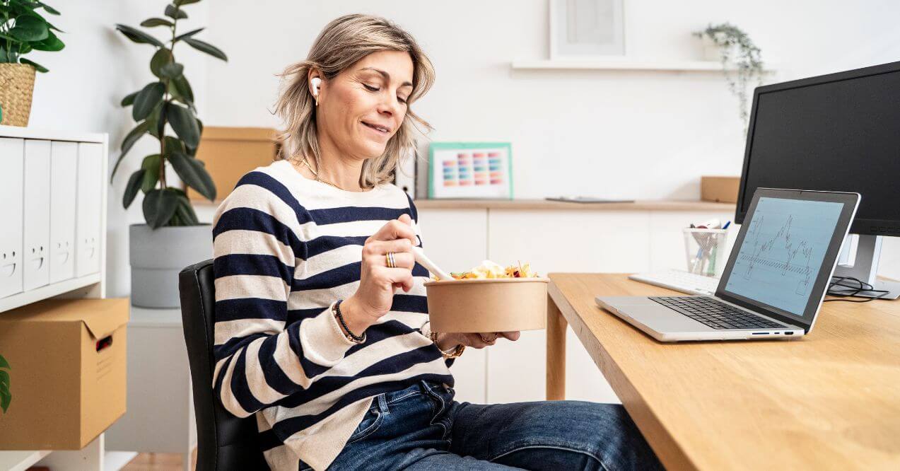 Woman eating mind salad from a bowl while working at her desk with a laptop.